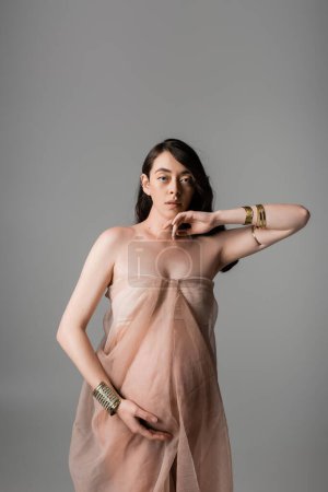 pretty and romantic mother-to-be looking at camera while posing in golden bracelets and airy chiffon draping isolated on grey background, maternity fashion concept, sensuality
