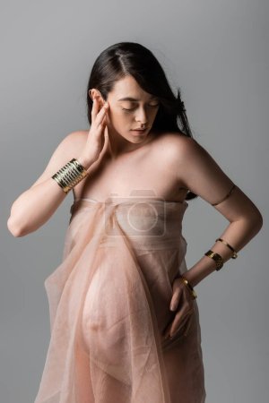 charming and elegant pregnant woman in golden bracelets and gentle chiffon draping posing with hand near face isolated on grey background, stylish pregnancy concept, sensuality