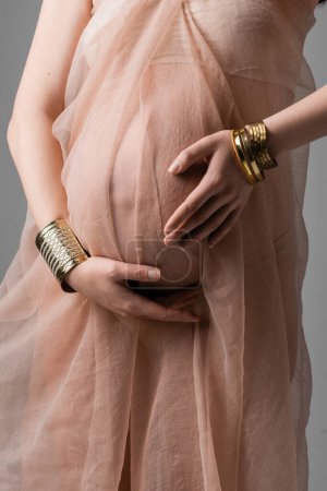 geschnittene Ansicht einer schwangeren Frau in sanftem Chiffontuch und goldenen Armbändern, die den Bauch isoliert auf grauem Hintergrund umarmen, Umstandsmodekonzept, zukünftige Mutter mit Bauch