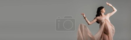 mujer embarazada elegante con cabello moreno ondulado posando en gasa fluida cortina y accesorios dorados aislados sobre fondo gris, concepto de embarazo elegante, mujer embarazada con vientre, pancarta