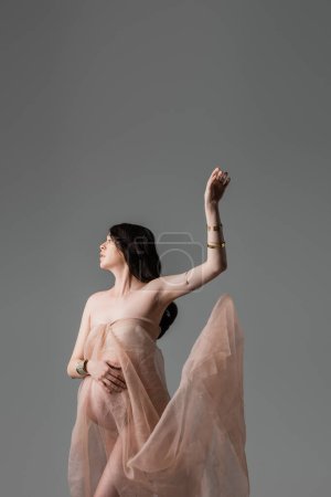romantische schwangere Frau in weichem und transparentem Chiffon-Drapierung und goldenen Accessoires posiert mit erhobener Hand isoliert auf grauem Hintergrund, Umstandsmodekonzept, Erwartung 