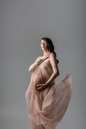 Foto de Mujer encantadora y embarazada en suave gasa drapeado y pulseras de oro sosteniendo la mano cerca del pecho aislado sobre fondo gris, concepto de estilo de maternidad, expectativa - Imagen libre de derechos