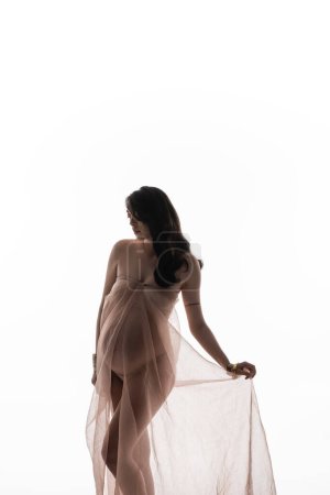silueta de elegante modelo embarazada con cabello castaño ondulado posando en tela de gasa transparente y aireada aislada sobre fondo blanco, concepto de moda de maternidad, expectativa 