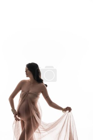 mujer atractiva y embarazada en pulseras doradas posando en gasa transparente y suave drapeado aislado sobre fondo blanco, concepto de maternidad elegante, expectativa, futura madre con vientre