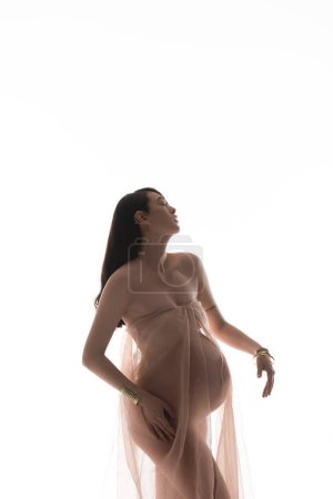 Charmante und romantische schwangere Frau posiert in goldenen Accessoires und transparentem Chiffondrapier isoliert auf weißem Hintergrund, Umstandsmodekonzept, Erwartung, zukünftige Mutter mit Bauch
