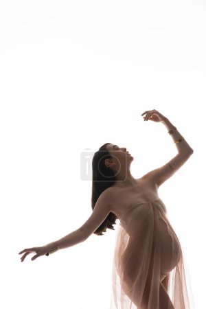 graceful mother-to-be in golden bracelets and delicate chiffon draping posing with closed eyes and raised hand isolated on white background, maternity fashion concept, pregnant woman 