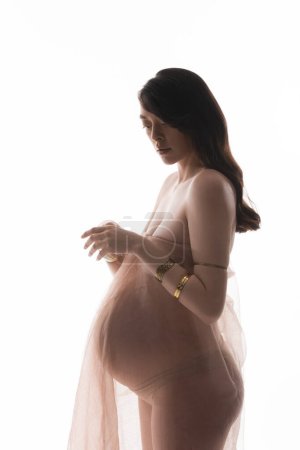 encantadora y elegante mujer embarazada morena en pulseras de oro y delicada gasa drapeado de pie aislado sobre fondo blanco, concepto de moda de maternidad, expectativa, futura madre con vientre