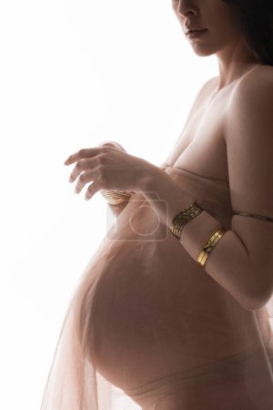 partial view of elegant expecting mother in golden accessories and transparent chiffon draping isolated on white background, maternity fashion concept, expectation, woman with belly