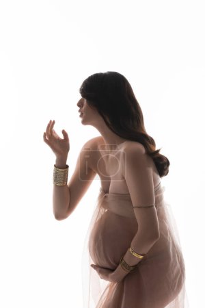 Foto de Vista lateral de la futura madre morena en pulseras doradas y tela de gasa delicada y transparente aislada sobre fondo blanco, concepto de moda de maternidad, expectativa, mujer con vientre - Imagen libre de derechos