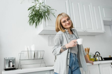 Photo for Happy young woman with short hair and bangs, eyeglasses and tattoo holding cup of morning coffee while standing in casual clothes next to toaster, dishes, kettle and plant in modern kitchen - Royalty Free Image