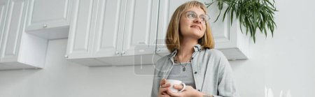 mujer joven positiva con pelo corto y flequillo, anteojos y tatuaje sosteniendo taza de café de la mañana mientras mira hacia otro lado y de pie en ropa casual al lado del gabinete de cocina blanco y la planta, pancarta 