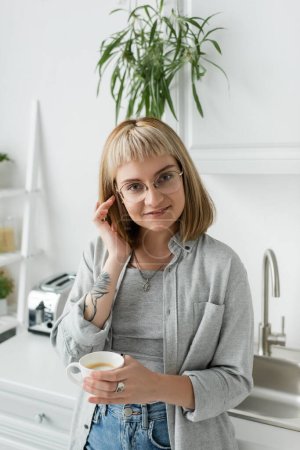 Photo for Happy young woman with bangs, eyeglasses and tattoo on hand adjusting short hair and holding cup of coffee while looking at camera, standing in casual clothes next to toaster, kitchen sink at home - Royalty Free Image