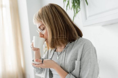 Photo for Young woman with bangs, eyeglasses and short hair holding cup while drinking morning coffee and standing in casual grey clothes next to blurred plant in kitchen - Royalty Free Image