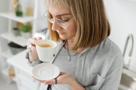 young woman with bangs, eyeglasses and short hair holding cup of morning coffee with saucer and standing in casual grey clothes next to blurred white wall in modern kitchen 