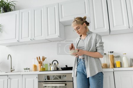 mujer joven con flequillo, anteojos y pelo corto sosteniendo tazón con desayuno y cuchara mientras está de pie en ropa gris casual al lado de hervidor de agua, electrodomésticos de cocina y armarios blancos borrosos en casa 