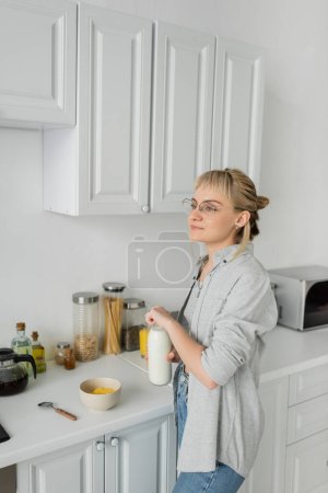 junge Frau mit Brille und kurzen Haaren mit Pony hält Flasche mit Milch in der Nähe Schüssel mit Cornflakes beim Frühstück und steht in lässiger Kleidung neben Küchengeräten zu Hause 