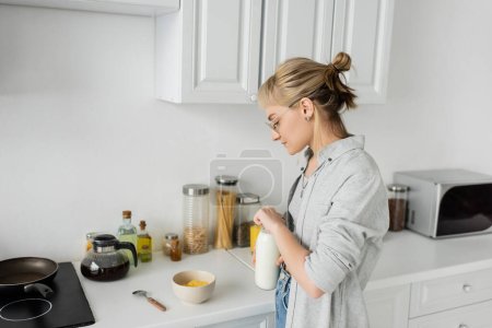 mujer joven en gafas graduadas y pelo corto con flequillo sosteniendo botella con leche fresca cerca de un tazón con copos de maíz mientras hace el desayuno y de pie en ropa casual al lado de los electrodomésticos de cocina en casa 