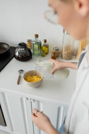 mujer joven borrosa y tatuada en gafas con botella mientras se vierte leche fresca en un tazón con copos de maíz en la encimera de la cocina mientras se hace el desayuno y de pie en la cocina moderna 