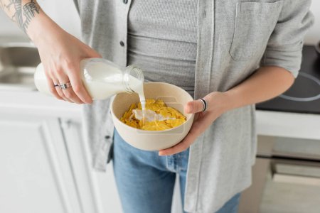 abgeschnittene Ansicht tätowierter junger Frau mit Flasche, während sie frische Milch in Schüssel mit Cornflakes gießt und in lässiger Kleidung mit Jeans in der modernen Küche frühstückt  