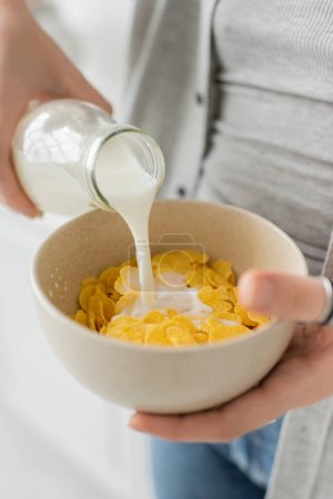 Nahaufnahme einer jungen Frau, die eine Flasche hält, während sie frische Milch in eine Schüssel mit Cornflakes gießt und in legerer Kleidung in der modernen Küche frühstückt  