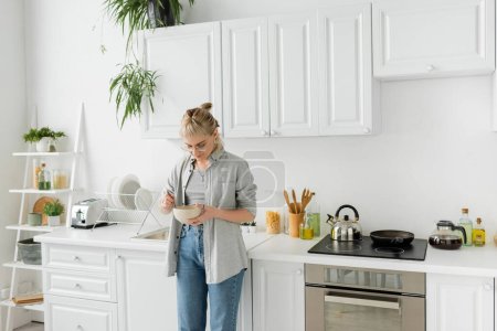 mujer joven con flequillo en anteojos sosteniendo tazón con copos de maíz y cuchara mientras está de pie en ropa gris casual y vaqueros al lado de los electrodomésticos de cocina en la cocina blanca borrosa en casa 