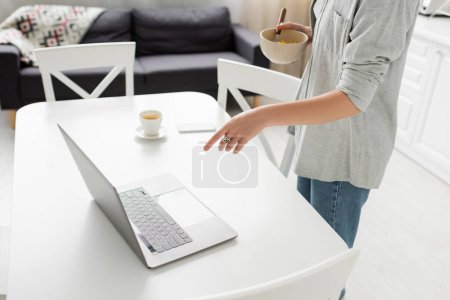 cropped view of young freelancer in casual outfit holding bowl with cornflakes while having breakfast and pointing at laptop near smartphone and cup of coffee on desk around chairs in modern kitchen 