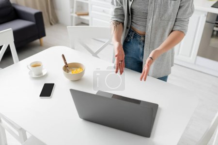 abgeschnittene Ansicht einer jungen Frau mit Tätowierung an der Hand, die auf Laptop neben Smartphone mit leerem Bildschirm zeigt, Schüssel mit Cornflakes, Löffel und Tasse Kaffee auf weißer Untertasse auf Schreibtisch in moderner Küche, freiberuflich