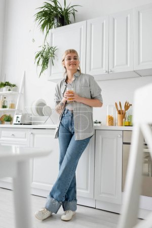 pleine longueur de femme tatouée et heureuse dans des lunettes tenant un verre de jus d'orange près du plan de travail de la cuisine avec vaisselle propre, grille-pain et rack avec des plantes regardant la caméra dans un appartement moderne 