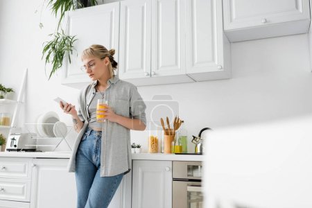 Tätowierte Frau mit Pony und Brille, die ein Glas Orangensaft in der Hand hält und das Smartphone benutzt, während sie neben sauberem Geschirr und grünen Pflanzen in verschwommener weißer Küche in einer modernen Wohnung steht 