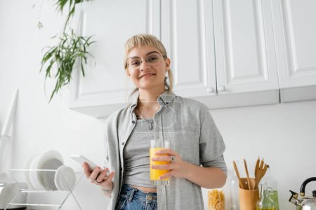 mujer tatuada y feliz con flequillo y anteojos sosteniendo vaso de jugo de naranja y teléfono inteligente mientras mira la cámara cerca de platos limpios y plantas verdes borrosas en apartamento moderno 