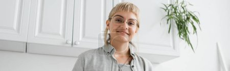 jeune femme heureuse avec une frange et des lunettes souriant tout en se tenant en tenue décontractée grise et regardant la caméra près des armoires de cuisine blanches et des plantes vertes floues dans un appartement moderne, bannière 