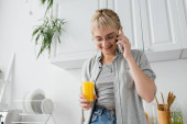 happy young woman with bangs and eyeglasses holding glass of orange juice and talking on smartphone standing with closed eyes in kitchen near blurred green plants in modern apartment  Mouse Pad 656114574