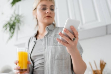 young woman in eyeglasses, with ring on finger holding glass of orange juice and smartphone while texting and standing in blurred white kitchen with green indoor plants in modern apartment 