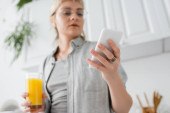 young woman in eyeglasses, with ring on finger holding glass of orange juice and smartphone while texting and standing in blurred white kitchen with green indoor plants in modern apartment  Poster #656114592