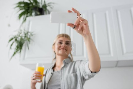 vista de ángulo bajo de la mujer feliz con flequillo y anillos en los dedos sosteniendo un vaso de jugo de naranja y tomando selfie en el teléfono inteligente y de pie en la cocina blanca borrosa con plantas verdes de interior 