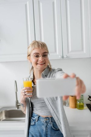 glückliche und tätowierte junge Frau mit Pony und Brille, die ein Glas Orangensaft in der Hand hält und ein Selfie mit verschwommenem Smartphone macht, während sie in der weißen Küche neben Spüle und Ölflasche steht