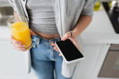 cropped view of young woman with tattoo on hand holding glass of fresh orange juice and smartphone with blank screen while standing in casual clothes with blue denim jeans in modern apartment  Poster #656114640