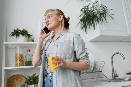 Foto de Mujer joven feliz con flequillo y anteojos sosteniendo un vaso de jugo de naranja y hablando en el teléfono inteligente, de pie cerca de las plantas verdes borrosas y estante en la cocina moderna y blanca - Imagen libre de derechos