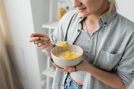 Photo for Cropped view of tattooed young woman smiling while holding bowl with cornflakes and spoon while having breakfast next to blurred white rack in modern kitchen. copy space, apartment, morning energy - Royalty Free Image