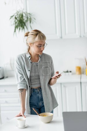 Photo for Young woman with short hair, tattoo and bangs using smartphone while standing in eyeglasses near bowl with cornflakes, cup of coffee and laptop on table in modern apartment, freelancer - Royalty Free Image