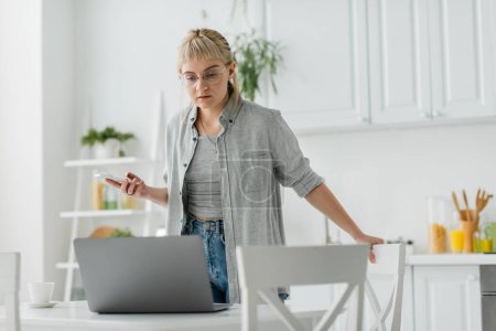 young woman with short hair, tattoo and bangs using smartphone while standing in eyeglasses near cup of coffee and looking at blurred laptop on table around chairs in modern apartment, freelancer 