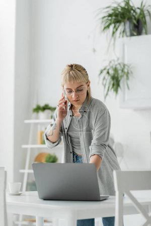 Photo for Young woman with short hair, bangs and tattoo on hand talking on smartphone while standing in eyeglasses near cup of coffee and laptop on table in modern apartment, freelancer - Royalty Free Image