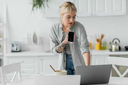 Photo for Young woman with bangs and tattoo on hand recording voice message on smartphone while standing in eyeglasses, bowl with cornflakes, cup of coffee and laptop on table in modern apartment, freelancer - Royalty Free Image