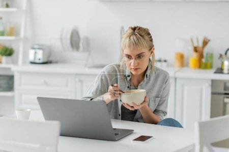 mujer joven con flequillo y tatuaje en la mano comer copos de maíz para el desayuno mientras se utiliza el ordenador portátil cerca de teléfono inteligente con pantalla en blanco y taza de café en la mesa en la cocina moderna, freelancer 
