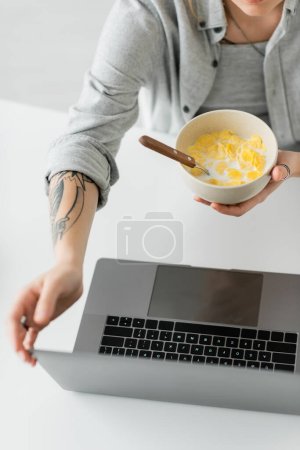 Foto de Vista recortada de mujer joven con tatuaje en la mano y camisa gris sosteniendo tazón con copos de maíz con cuchara mientras desayuna y el uso de la computadora portátil en la mesa blanca en el apartamento moderno, freelancer, vista superior - Imagen libre de derechos