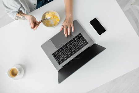 vue du dessus de la jeune femme avec tatouage sur la main manger des flocons de maïs pour le petit déjeuner tout en utilisant un ordinateur portable près du smartphone avec écran blanc et tasse de café sur la table dans la cuisine moderne, pigiste, plan recadré