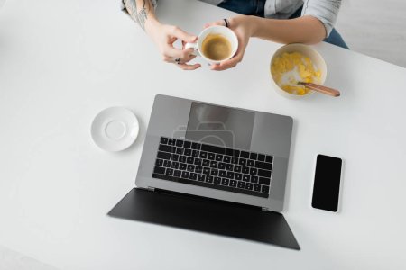 top view of woman with tattoo on hand holding cup of coffee near bowl with cornflakes during breakfast while using laptop near smartphone with blank screen in modern kitchen, freelancer, cropped 