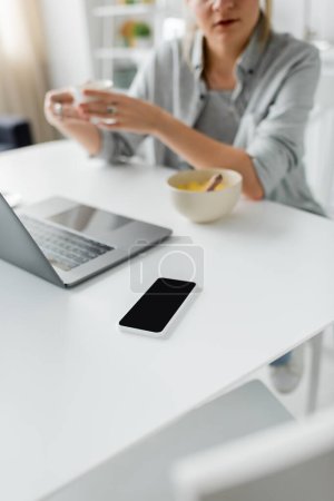 abgeschnittene Aufnahme einer verschwommenen Frau, die während des Frühstücks eine Tasse Kaffee in der Nähe einer Schüssel mit Cornflakes hält, während sie Laptop in der Nähe eines Smartphones mit leerem Bildschirm in der modernen Küche benutzt, Freelancer 