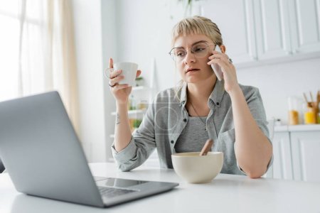 young woman with tattoo on hand and bangs talking on smartphone while using laptop, holding cup of coffee near bowl wth cornflakes on table in modern kitchen, freelancer, work from home 