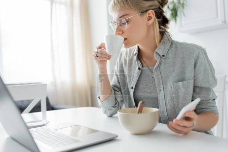 mujer joven con tatuaje en la mano y flequillo que sostiene el teléfono inteligente y el uso de la computadora portátil, beber café mientras sostiene la taza cerca y bowl wth copos de maíz en la mesa en la cocina moderna, freelancer 